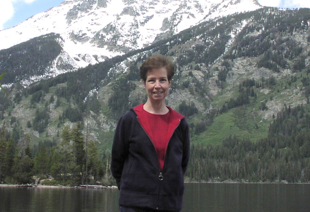 Linda Walker smiling, standing on the shore of an alpine lake with snow-capped mountains behind her.